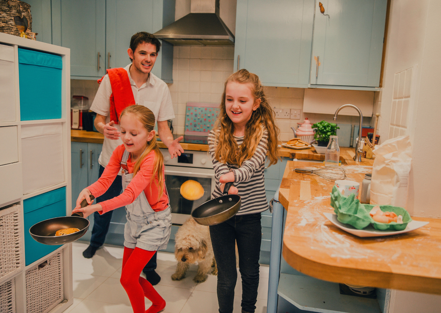 Image shows two young girls flip pancakes in a lively kitchen as a man watches with a smile. A small dog stands nearby, and baking ingredients are scattered on the counter.